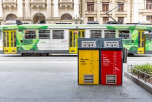 Yellow recycling and red waste PEL solar compacting bins on a Melbourne CBD footpath, with a green tram passing behind