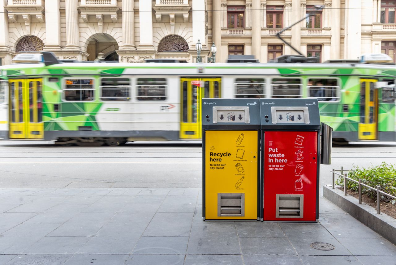 Yellow recycling and red waste PEL solar compacting bins on a Melbourne CBD footpath, with a green tram passing behind
