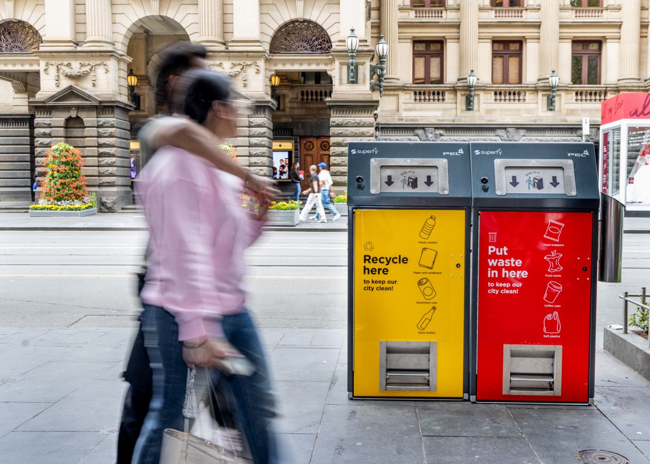 Twin yellow recycling and red waste bins with Superfy and PEL logos beside a stone building as pedestrians walk past